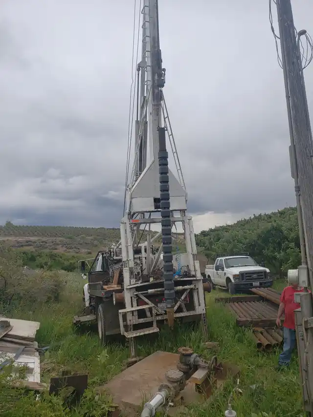 An Akland pump rig with its mast raised at a well in open scrub land under a grey sky, a pickup parked behind