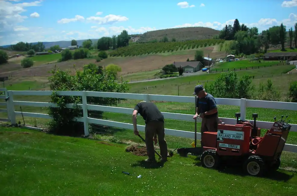 Two Akland crew members trenching a lawn with a ride-on trencher labelled AKLAND PUMP, a white fence and orchard hills behind