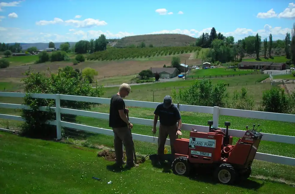 Two Akland crew members standing beside their AKLAND PUMP trencher on a lawn above the valley