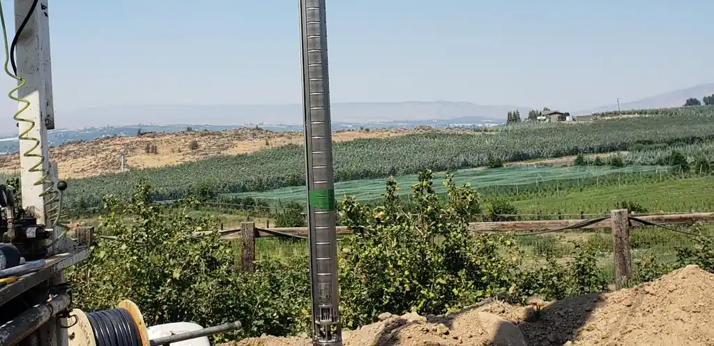 A stainless submersible pump column hanging from Akland's rig on a hillside above orchard rows and the Yakima Valley