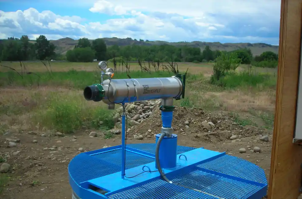 A blue steel grate platform over a well with a stainless discharge assembly, a field and hills behind