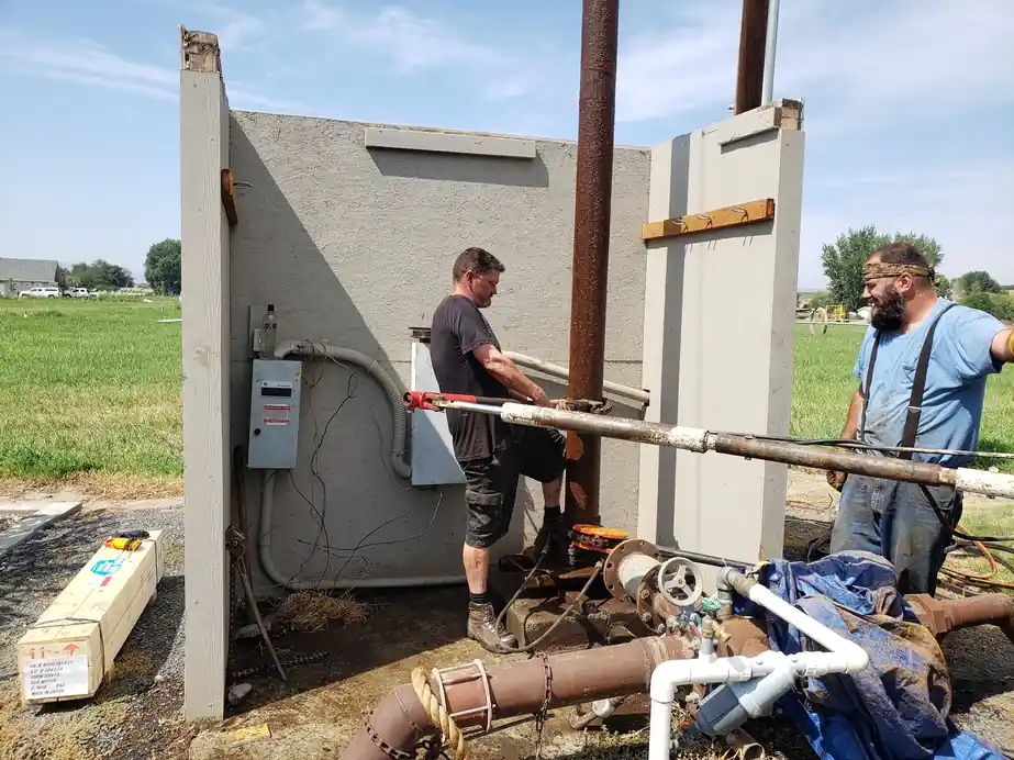 Two Akland crew working at a concrete pump house wall with a panel and piping, field behind them